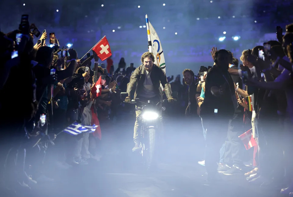 Chris Pratt rides a motorcycle with Olympic and Swiss flags, surrounded by cheering fans taking photos