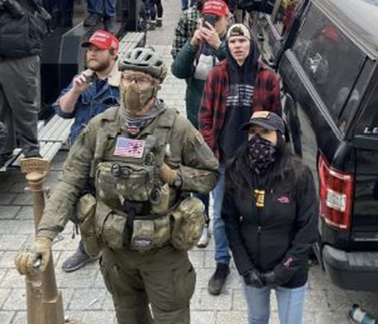 Jeremy Brown, in khaki-colored military fatigues and bicycle helmet, waits with others at the rally, including a short woman in a baseball cap, and two men in MAGA red baseball caps, outside the Capitol.