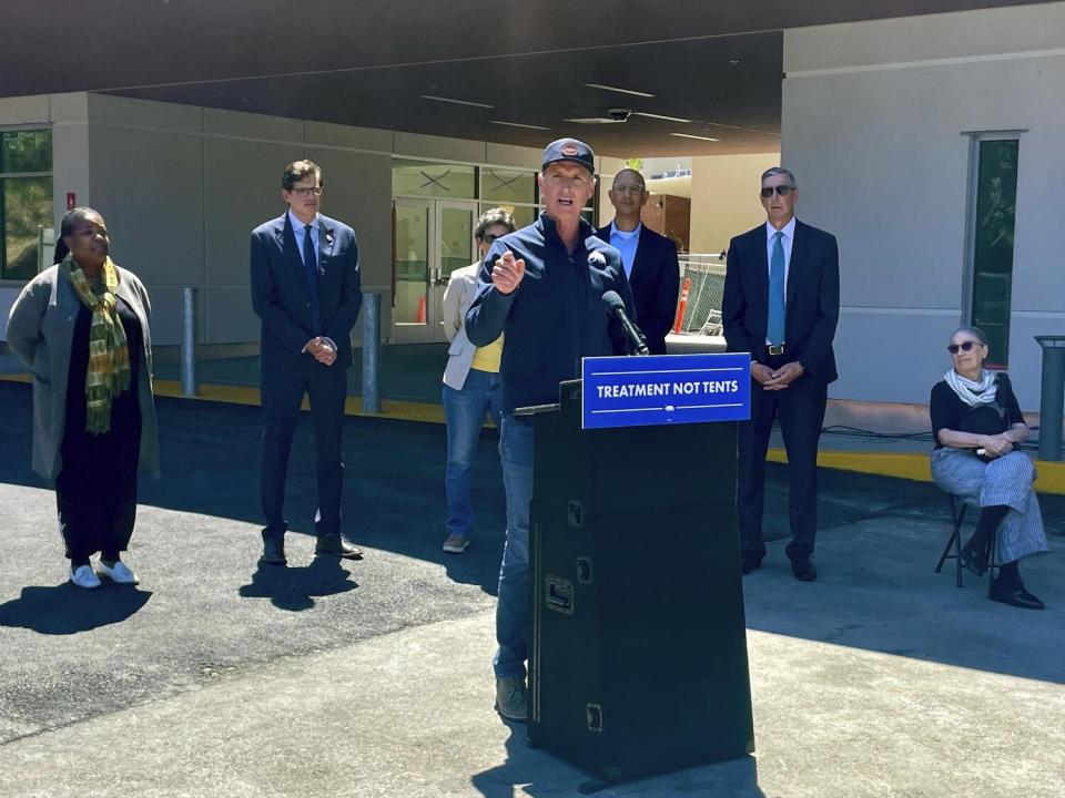 Gov. Gavin Newsom, in a blue shirt and baseball cap, speaks at a lectern.