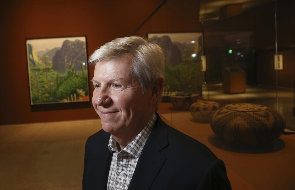 In this Wednesday, Oct. 23, 2019, photo, Richard Benefield, former executive director of the David Hockney Foundation, smiles as he looks inside the exhibit of Hockney's Yosemite artistic work, background, along with baskets from weavers from the Miwok and Mono Lake Paiute tribes on display at the Heard Museum in Phoenix. "David Hockney's Yosemite and Masters of California Basketry" exhibition opens Monday. (AP Photo/Ross D. Franklin)