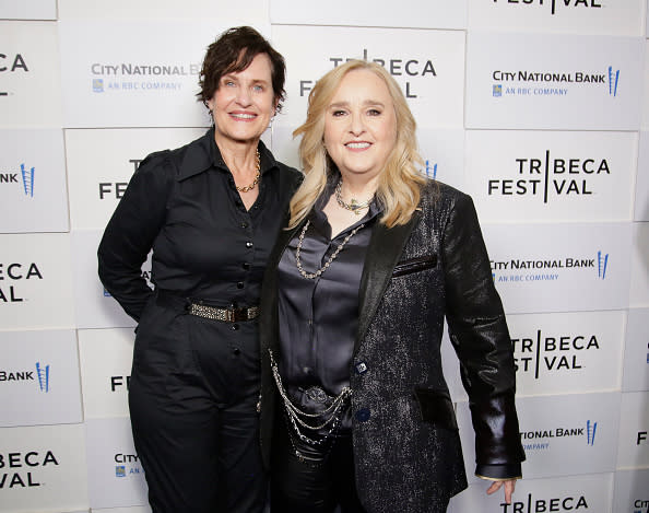 Linda Wallem and Melissa Etheridge pose together at the Tribeca Festival backdrop. Linda wears a belted jumpsuit, and Melissa sports a shiny blazer over a satin shirt