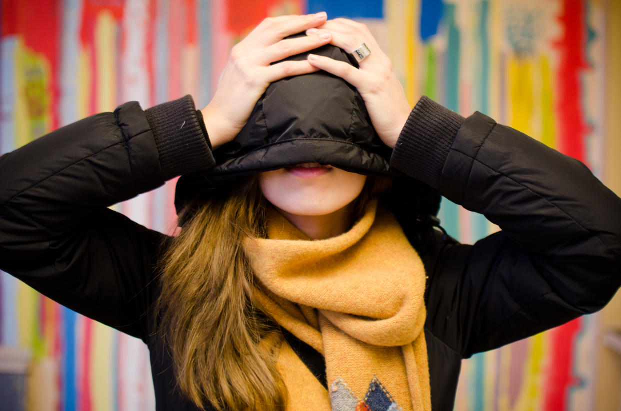 Beautiful young woman hiding face behind hoodie. In black jacket with scarf.