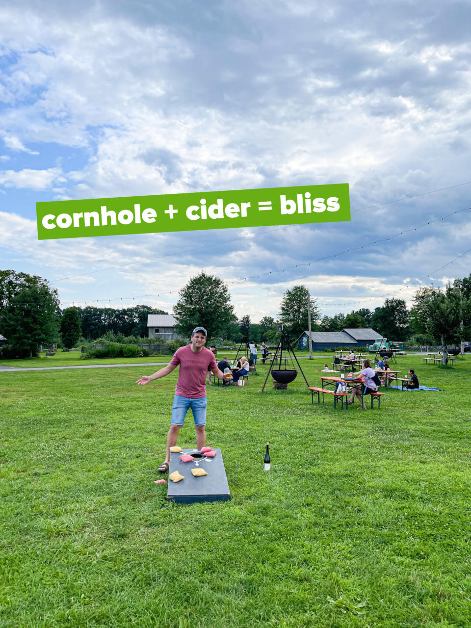 A man standing amid a field with people at outdoor tables and a fire pit with the text "cornhole + cider = bliss"