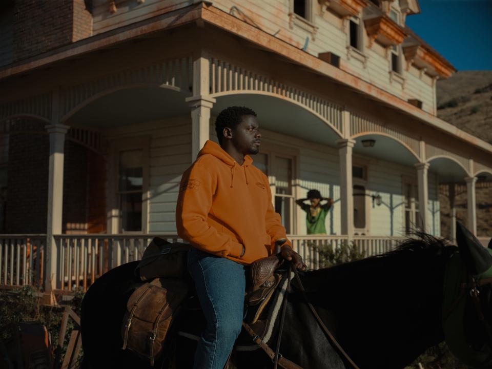 OJ sits astride a horse in an orange hoodie, while Emerald stands on the porch of their home behind him in a green jersey.