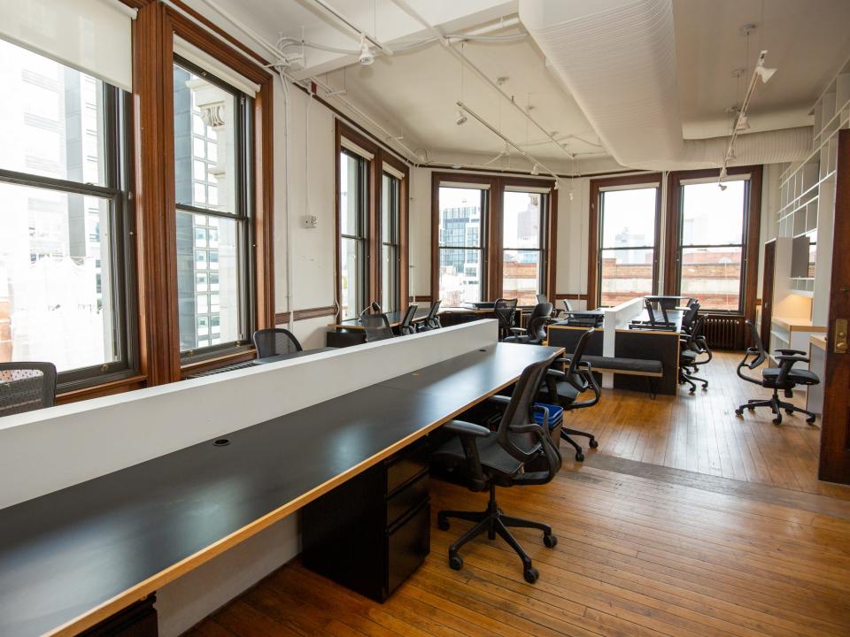Long desks with office chairs, shelves on the wall, and windows with views of nearby buildings.