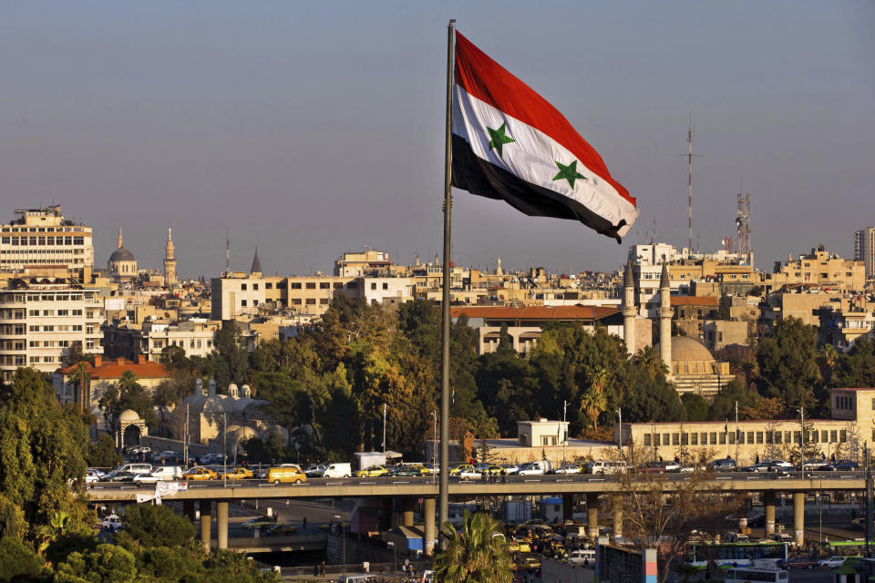 FILE - In this Feb. 28, 2016 file photo a Syrian national flag waves as vehicles move slowly on a bridge during rush hour, in Damascus, Syria. A new Syrian law empowering the government to confiscate property is threatening to leave refugees stuck in Europe with no homes to return to. (AP Photo/Hassan Ammar, file)