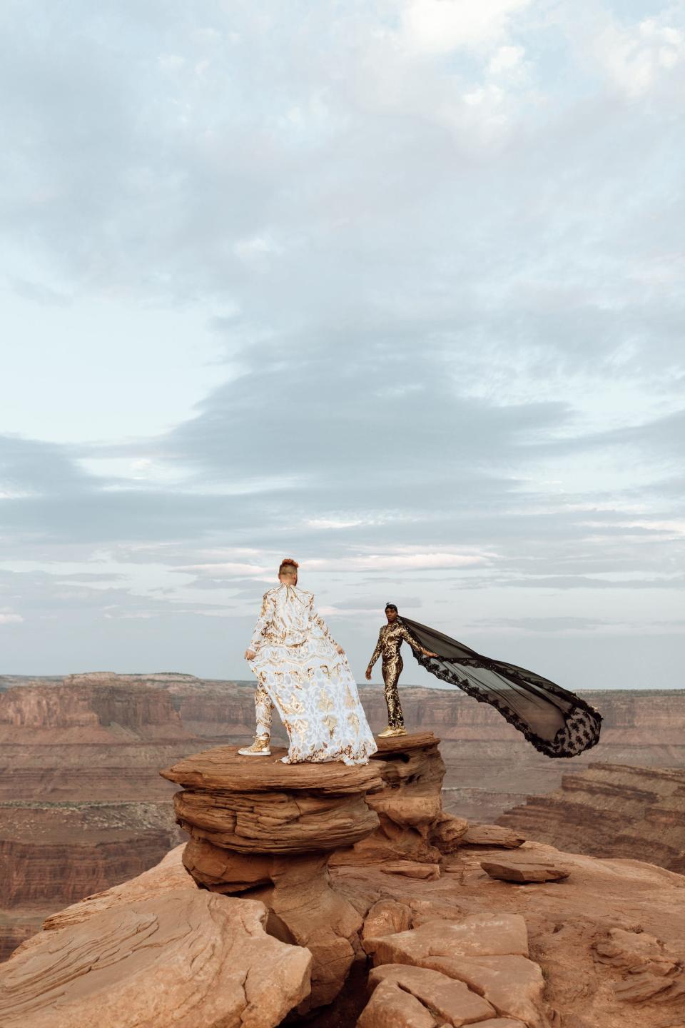 Two grooms stand in their wedding attire on a rock.