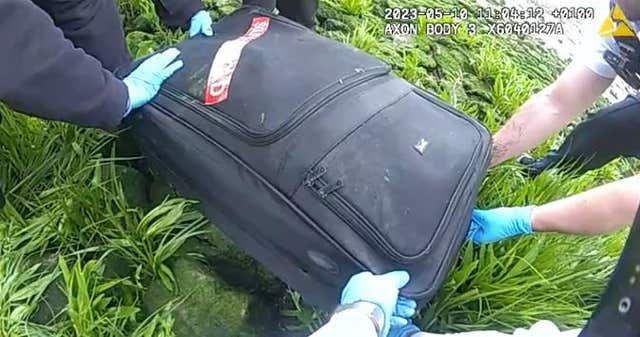 Hands with blue plastic gloves lifting a black suitcase from grassy area