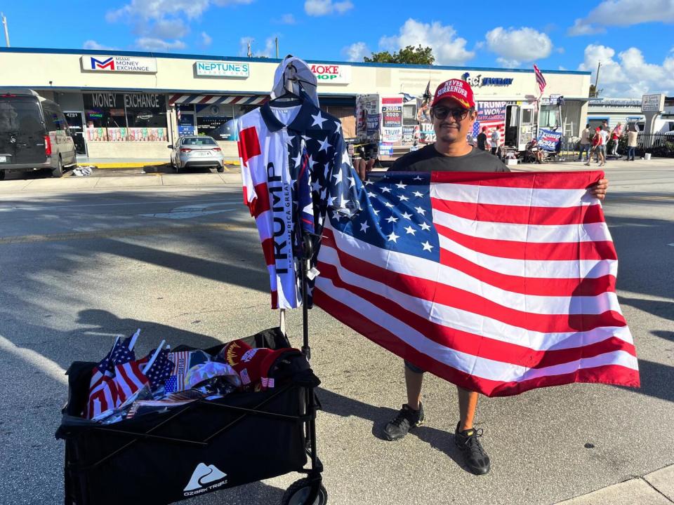 A man stands in a shopping center parking lot holding a large American flag by a wagon full of flags and Trump merchandise