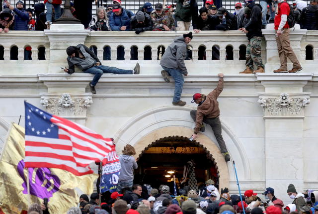Supporters of U.S. President Donald Trump climb over a portico onto a balcony as other rioters wearing red baseball caps and carrying a large American flag wait below.