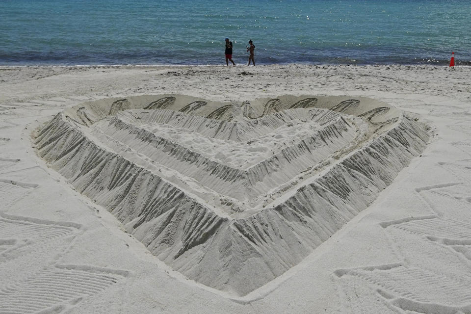 A giant heart sculpted from sand adorns the beach beside the area that is closed for search and rescue operations at the partially collapsed Champlain Towers South condo building, Friday, July 2, 2021, in Surfside, Fla. A crane works at the Champlain Towers South site in the distance, while Champlain Towers North is seen at right, and Champlain Towers East is seen center right. (AP Photo/Gerald Herbert)