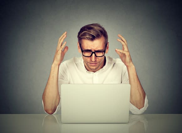 A man raises his hands in despair as he looks at the monitor on a laptop.