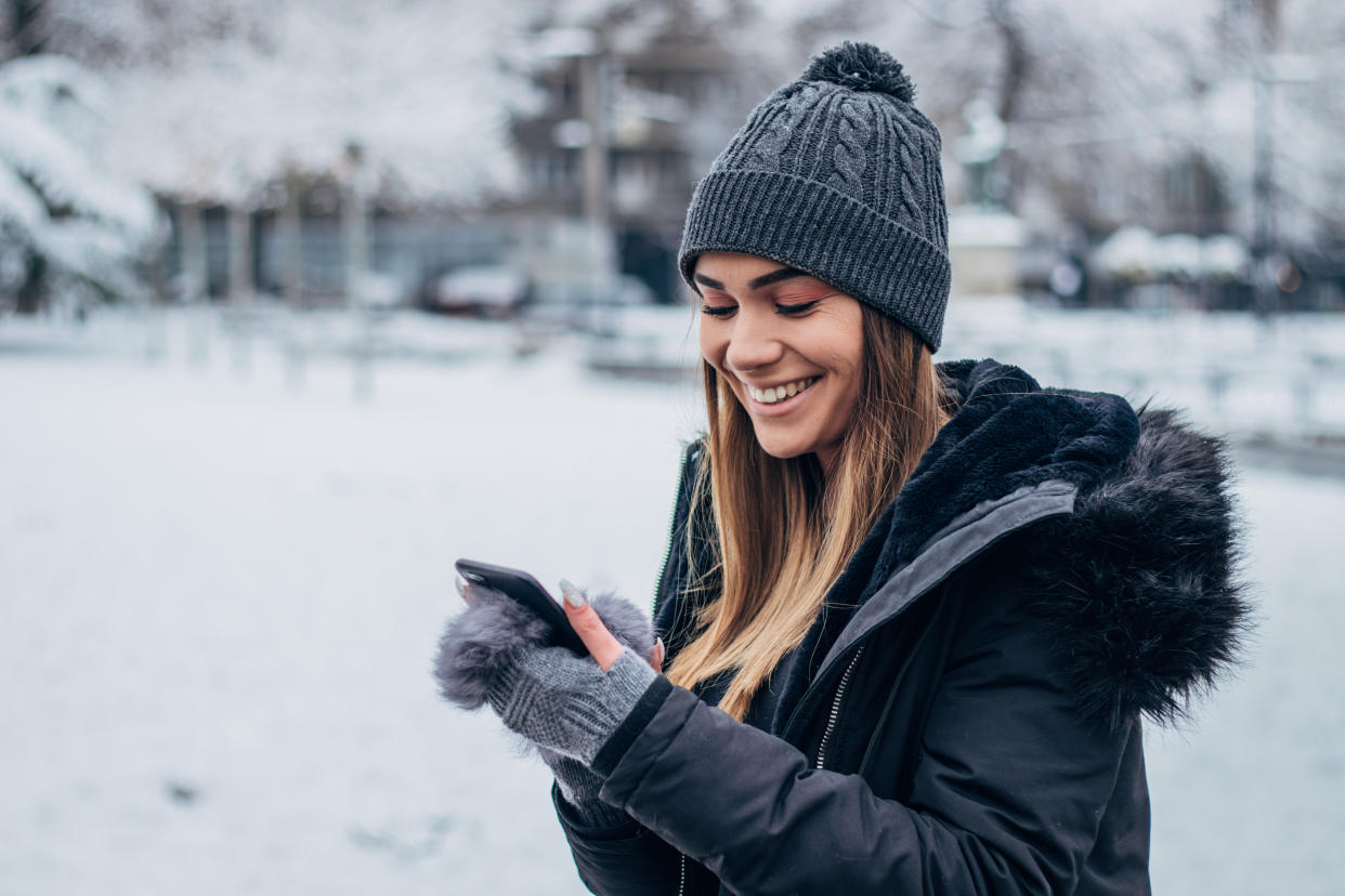 Beautiful young woman in winter clothing standing in the park covered with snow and text messaging.