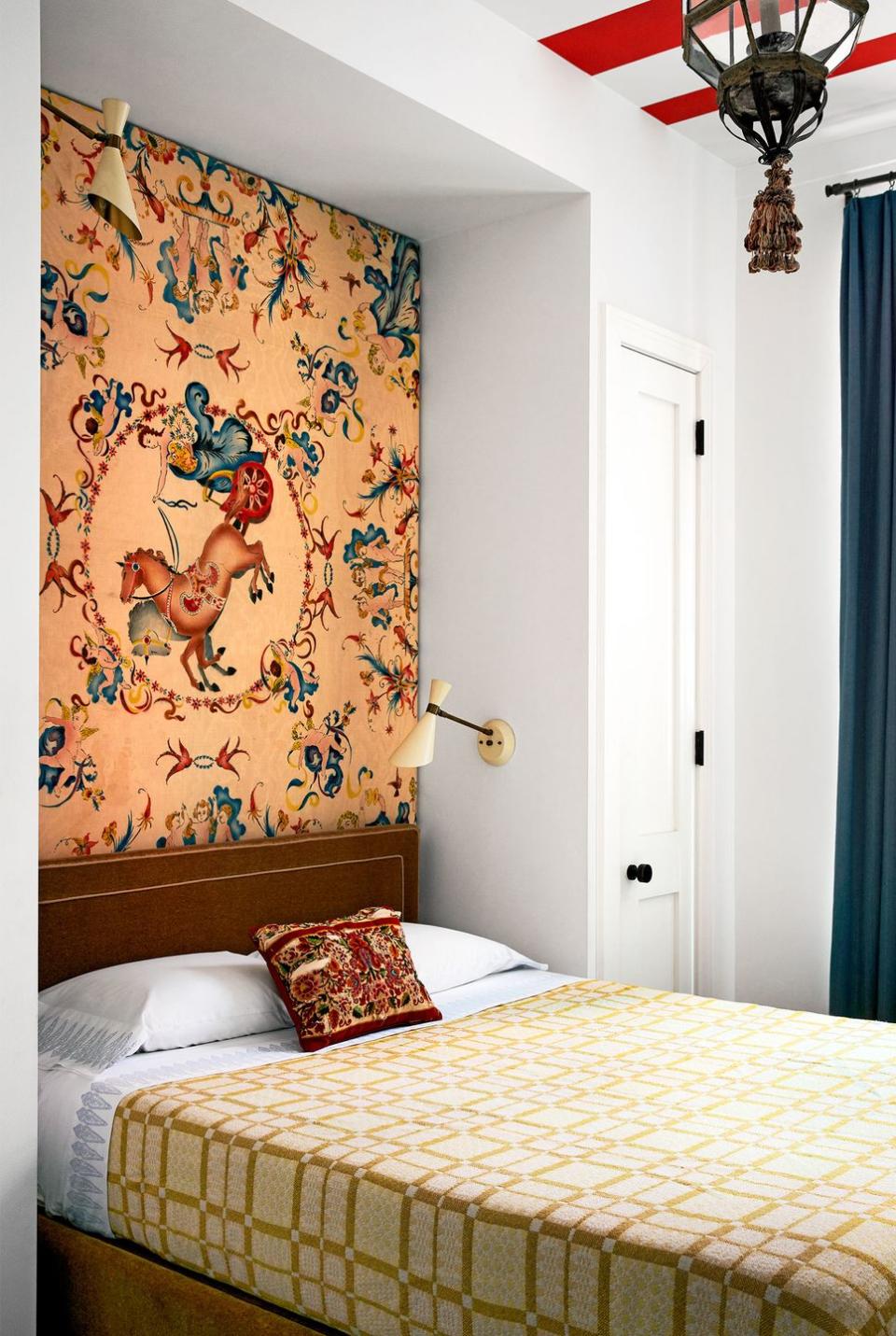 twin bed with golden yellow coverlet with rectangular pattern and a highly patterned wallpaper with a dancing horse and carriage at center over the head of the bed