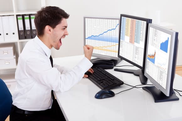A cheering man looking at stock charts on three computer screens and pumping his right fist.