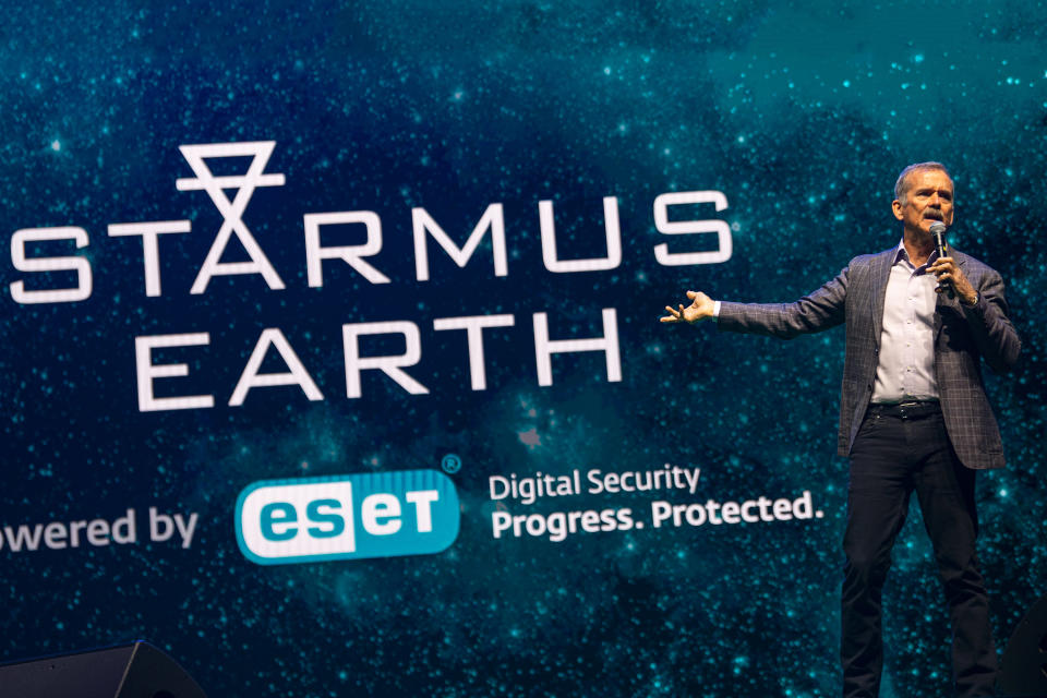 Ein Mann steht mit einem Mikrofon in der Hand vor einem großen Bildschirm, auf dem in großen Buchstaben die Worte „Starmus Earth“ geschrieben sind.