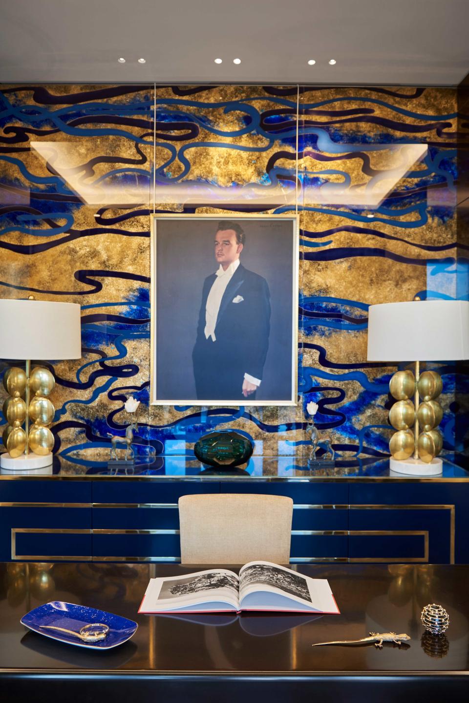 In an office, a portrait of Prince Rainier III as a young man is framed by a hand-painted glass wall in gold and navy hues.