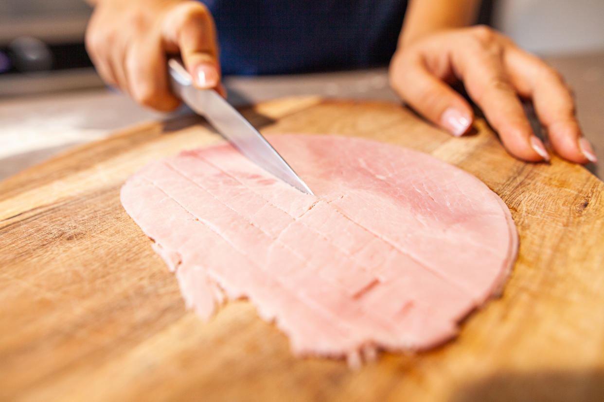 Une personne en train de découper des morceaux de jambon sur une planche en bois à l'aide d'un couteau (crédit : getty image)