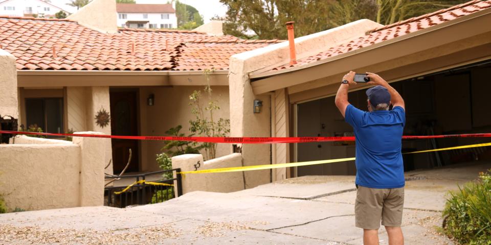 man taking photo of red-roof home, which has a crack in it and is behind red and yellow tape