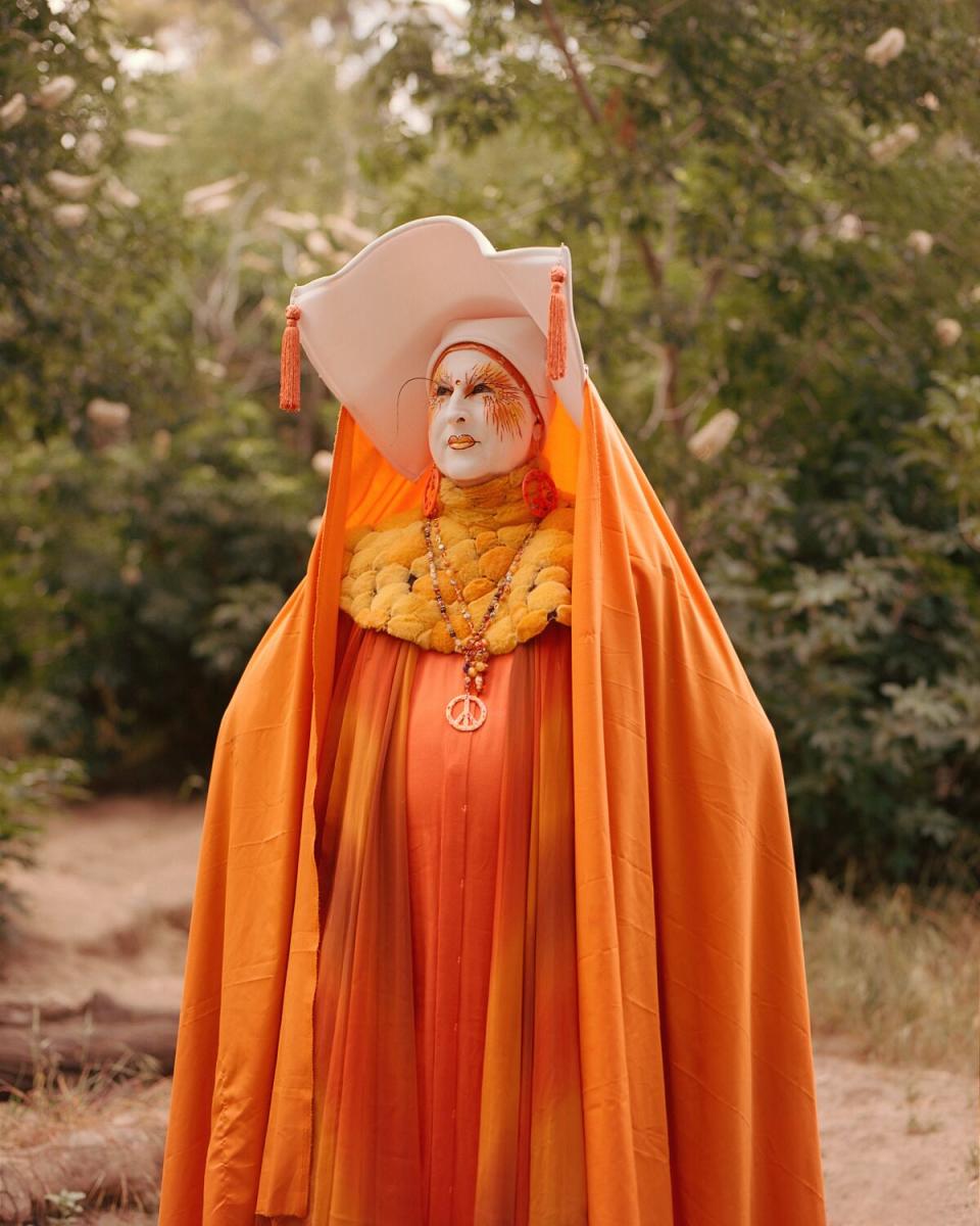 A drag nun in white face paint, wimpole and elaborate orange dress and cape.
