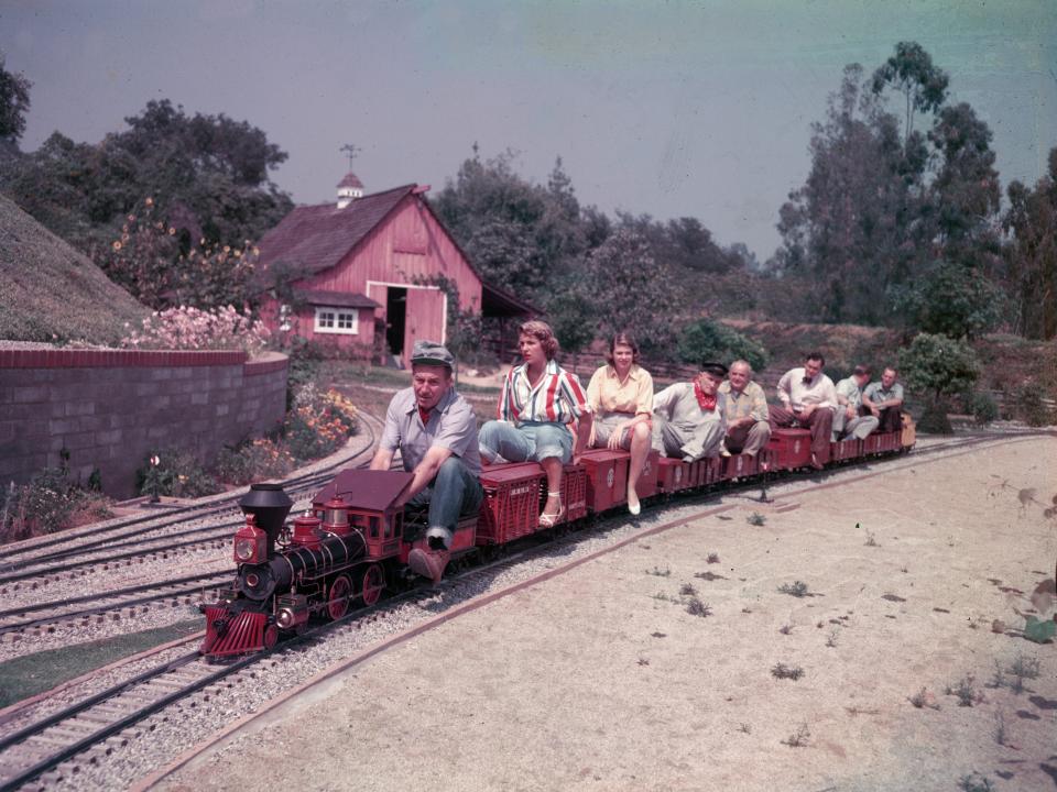 Walt Disney drives a miniature railroad with a line of passengers behind him at his home.