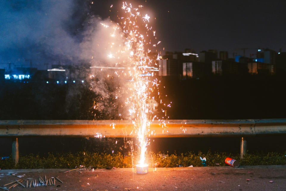 Fireworks spark and light up near a road at night, with buildings visible in the background