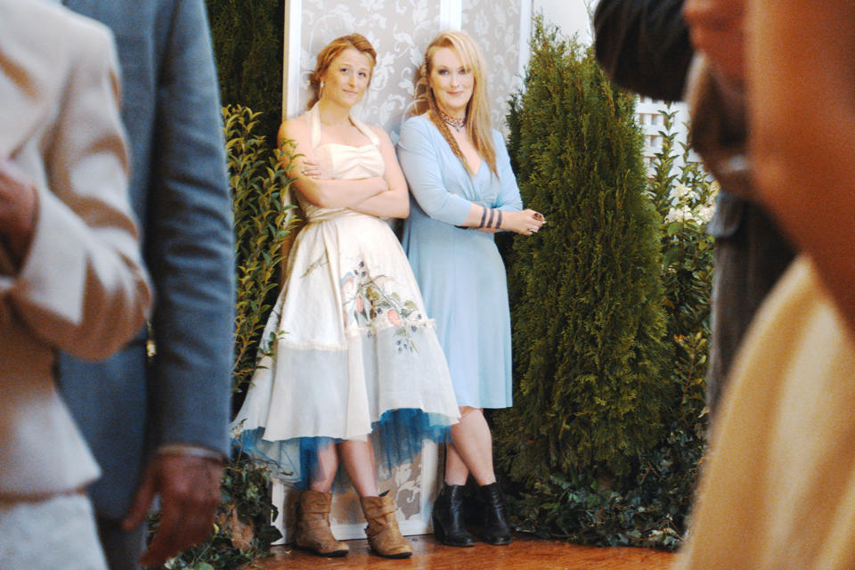 Two characters from a TV show stand together, one in a floral dress and cardigan, the other in a blue dress and boots