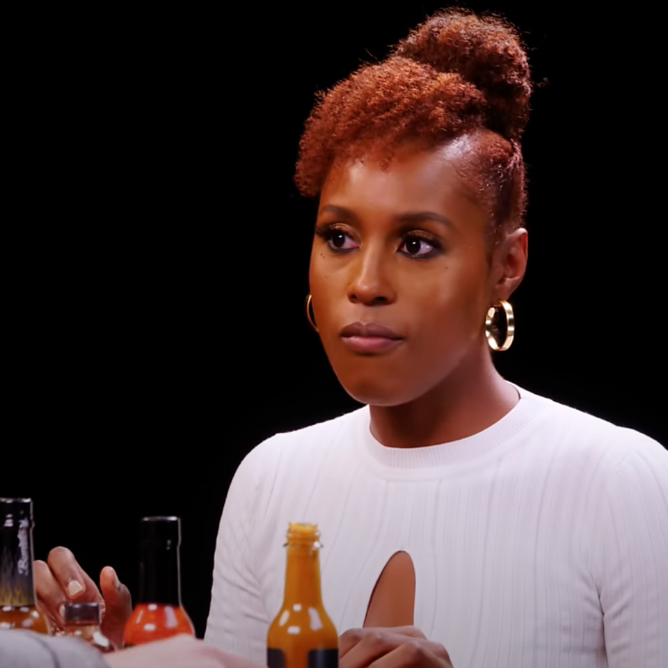 Issa Rae sits at a table during a recording, with various hot sauce bottles in front of her. She is wearing a white top with a cutout detail and large hoop earrings