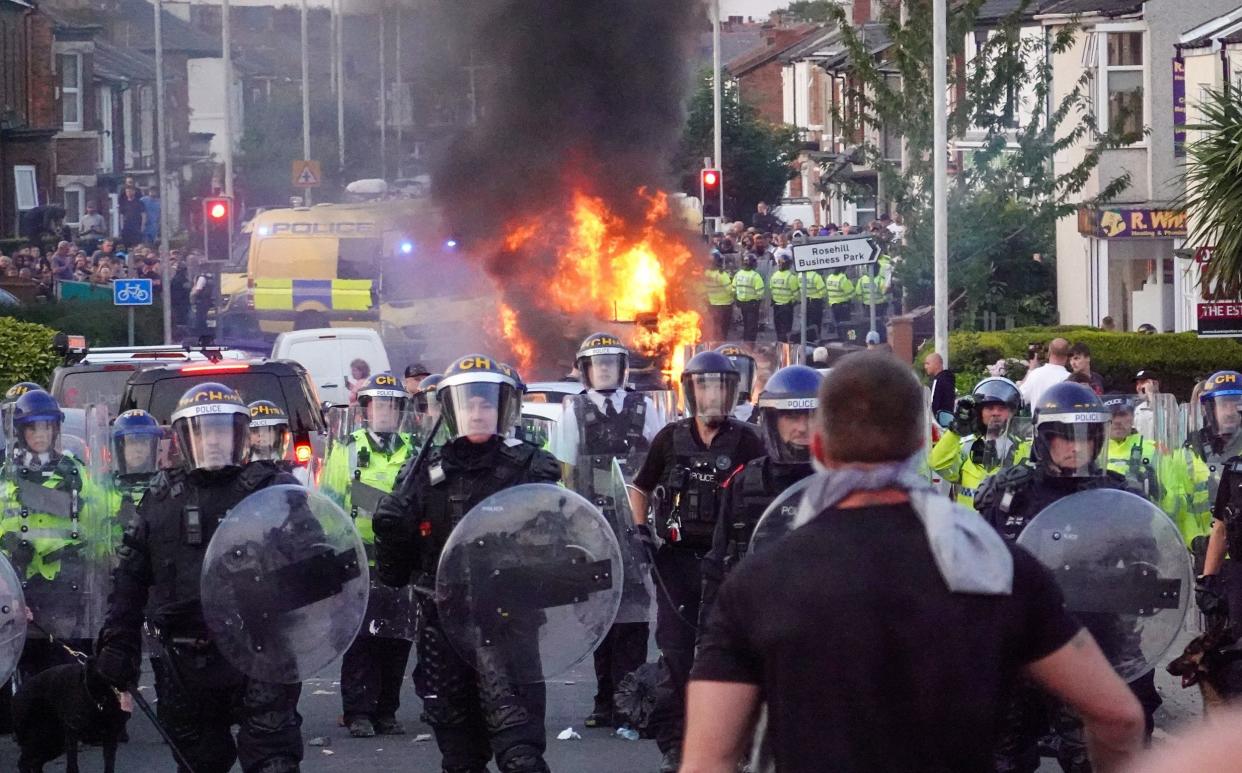 Riot police hold back protesters near a burning police vehicle