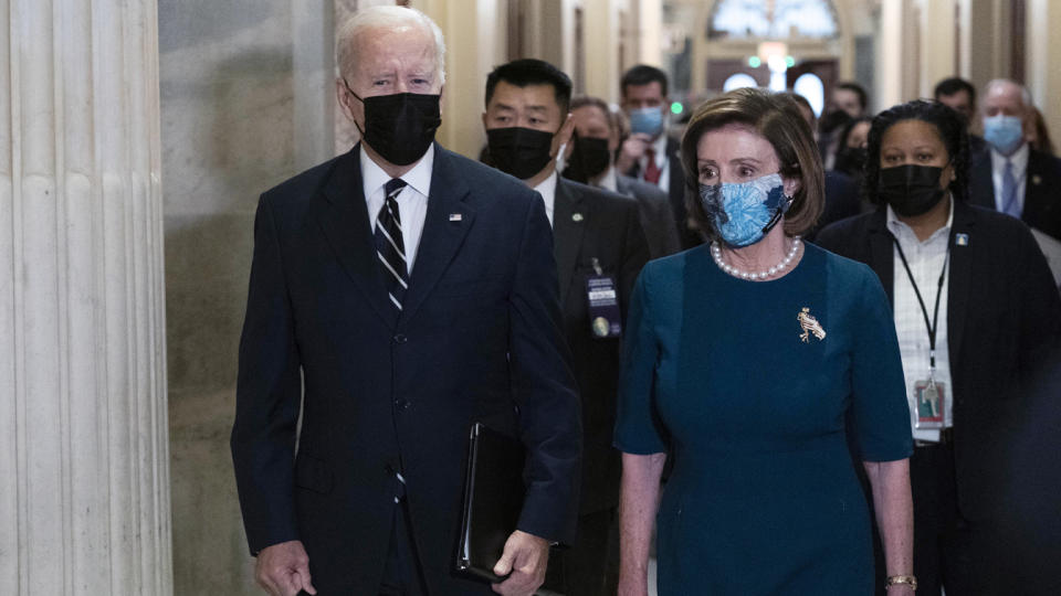 President Biden walks with Speaker of the House Nancy Pelosi in a hallway, while others trail behind them, all wearing face masks.