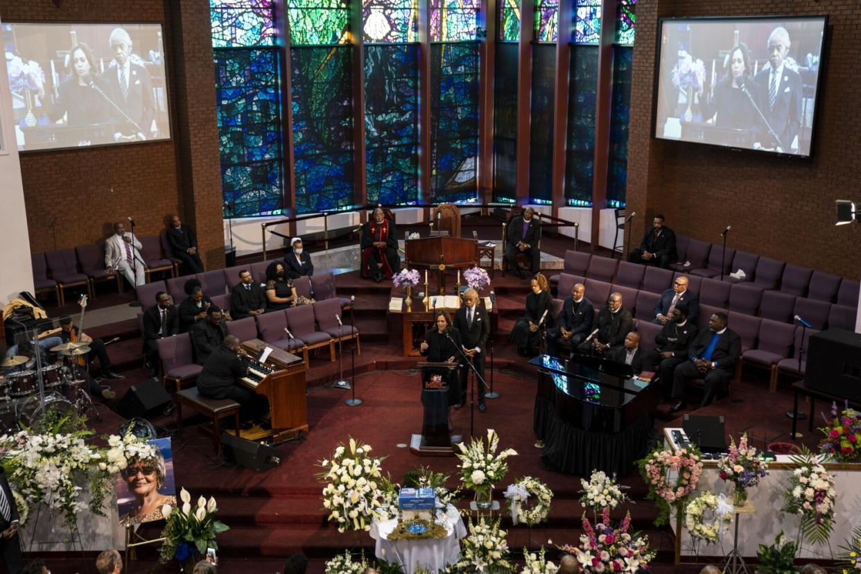 Kamala Harris speaks at funeral in a church with many funeral wreaths and people in seats.