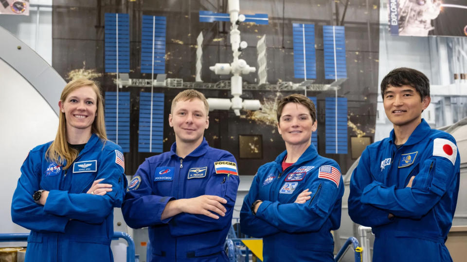  Two men and two women in blue flight suits stand in front of a model of the international space station. 