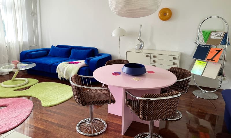 A pink tabling in a white-walled living room with four chairs and a book display stand.