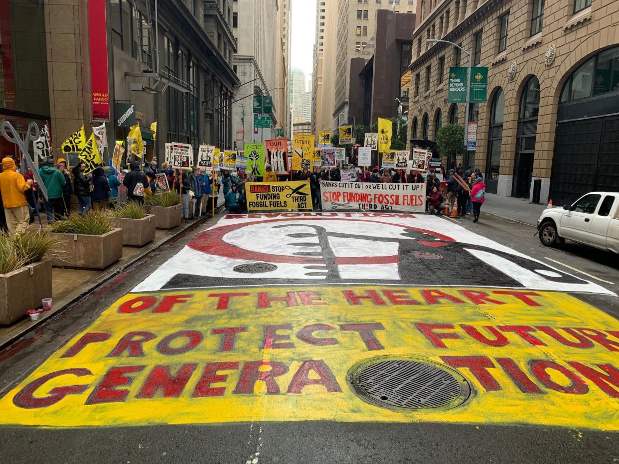 crowd with protest signs gathers around street painted yellow and red letters reading revolution of the heart protect future generations