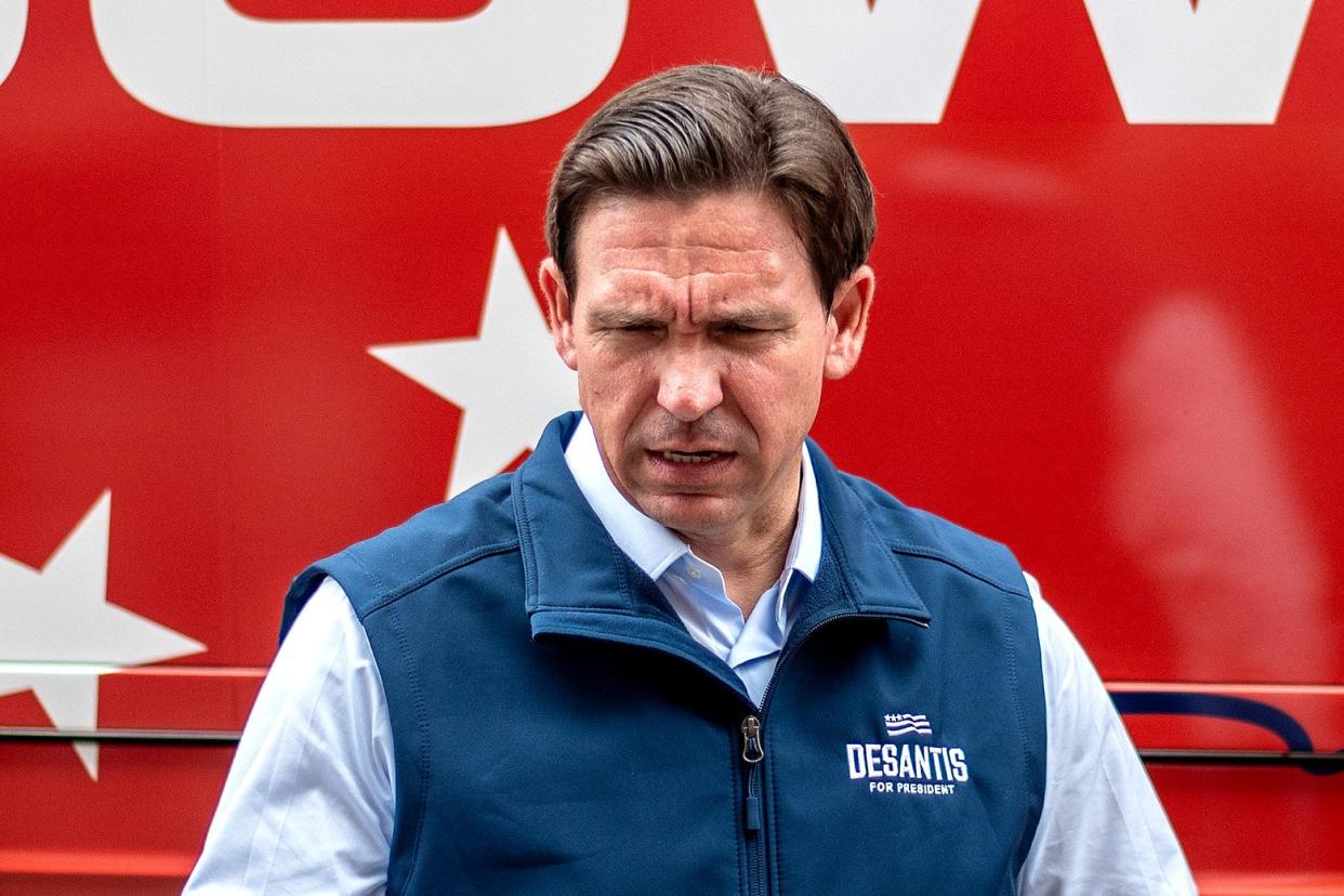 DeSantis looks downward, his brow furrowed, in front of his bus, wearing a DeSantis for President fleece vest