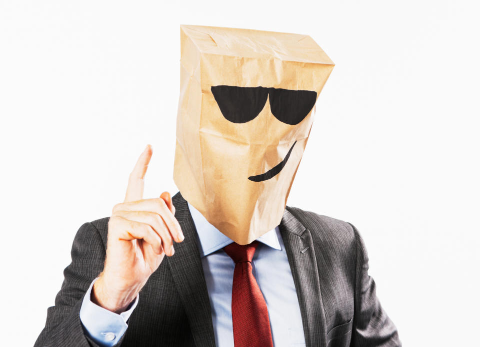 Person with a paper bag over head, smiling face drawn on it, suit-clad, raising index finger