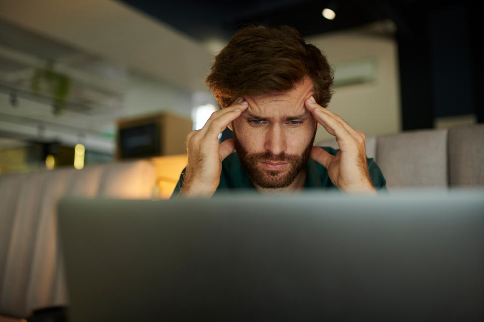 A man is seated looking at his laptop with a stressed expression, hands on his temples. The background suggests a modern indoor setting