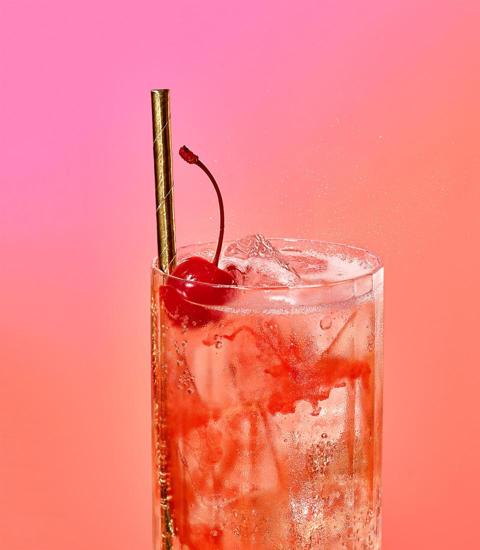 A soda in a glass with a cherry and straw on a bright pink background