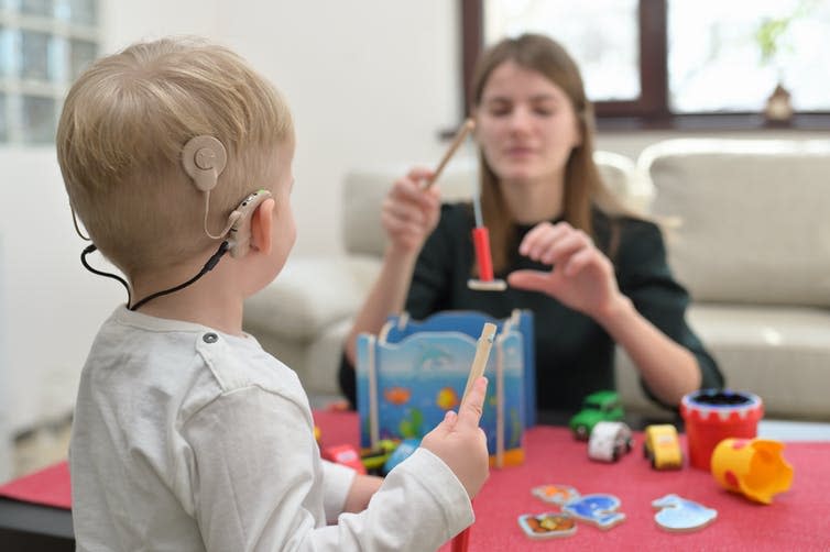 A young child wears a cochlear implact while playing with toys, sitting across from an adult.