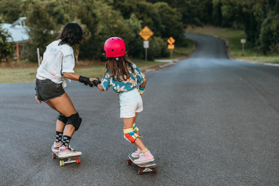 Two people skateboarding on a road: an adult and a child in protective gear, holding hands. Road signs and greenery in the background