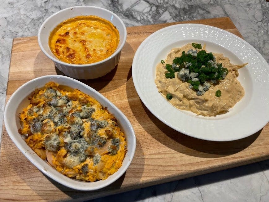 Cutting board with three bowls and plates of Buffalo-chicken dips. One bowl has a dip with crumbled blue cheese on top, one bowl has a dip with melted yellow cheese, and one dip is topped with crumbled blue cheese and scallions