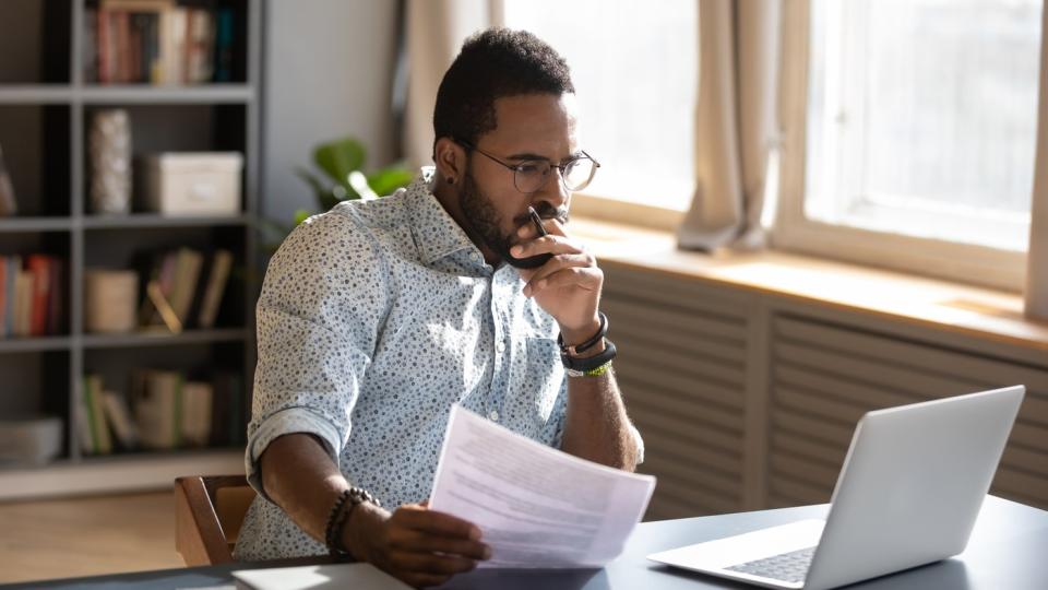 An investor looks pensively at a laptop while holding some papers.