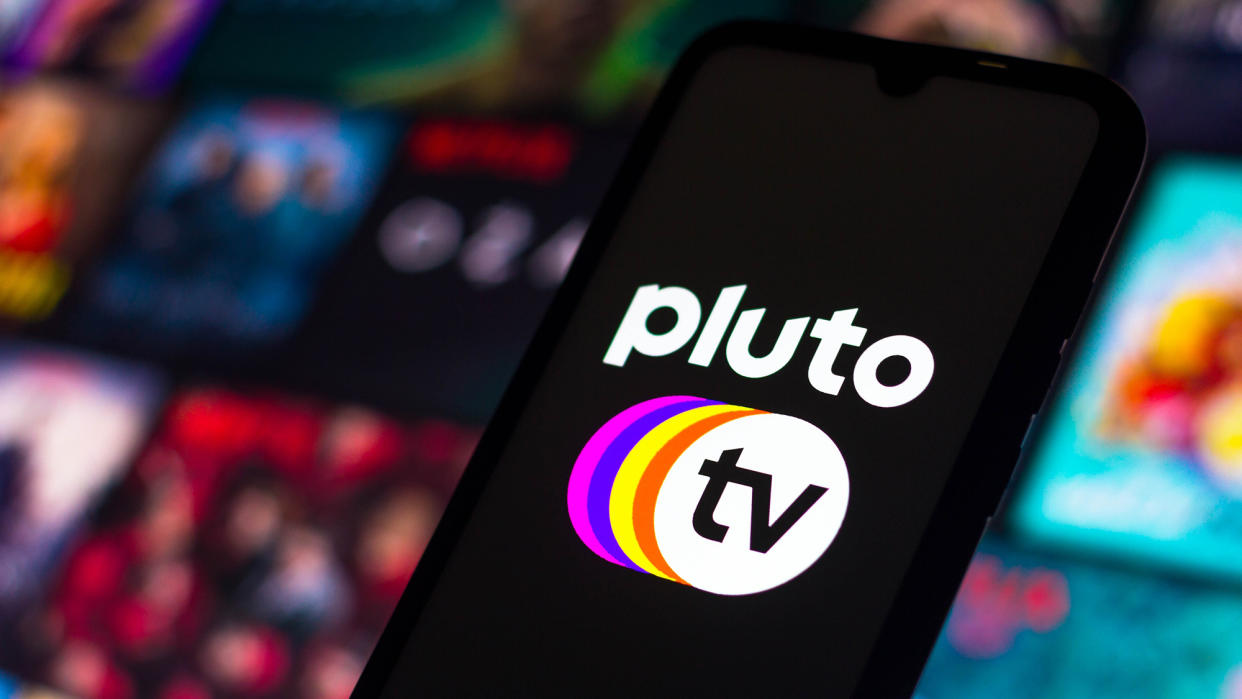  Pluto TV logo seen on a phone in front of a screen of blurry TV show images 