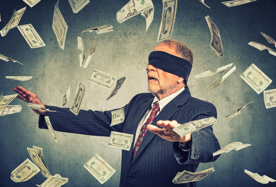 A blindfolded businessman chasing twenty-dollar bills floating in the air around him.