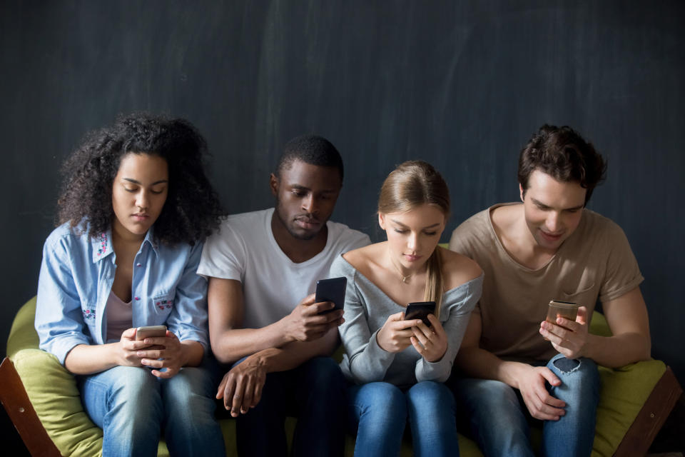 Young serious addicted to technology mixed race people sitting on couch in row, ignoring each other, using applications in smartphones, surfing websites, typing messages, scrolling social networks.