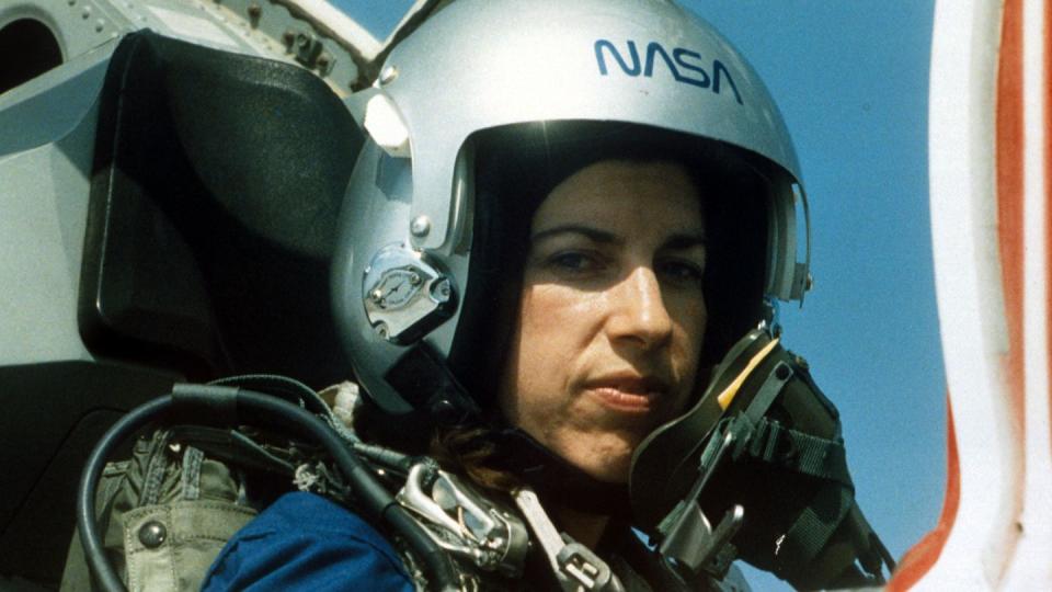 ellen ochoa sitting in an airplane cockpit and wearing a helmet while looking at the camera