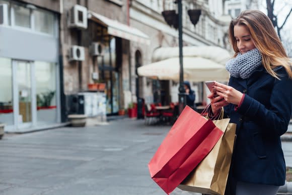 A woman looks at her phone while carrying shopping bags.