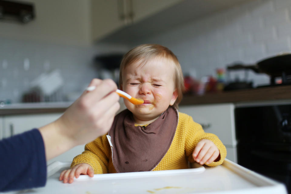 Toddler grimacing while being fed with a spoon by an adult's hand, seated in a high chair
