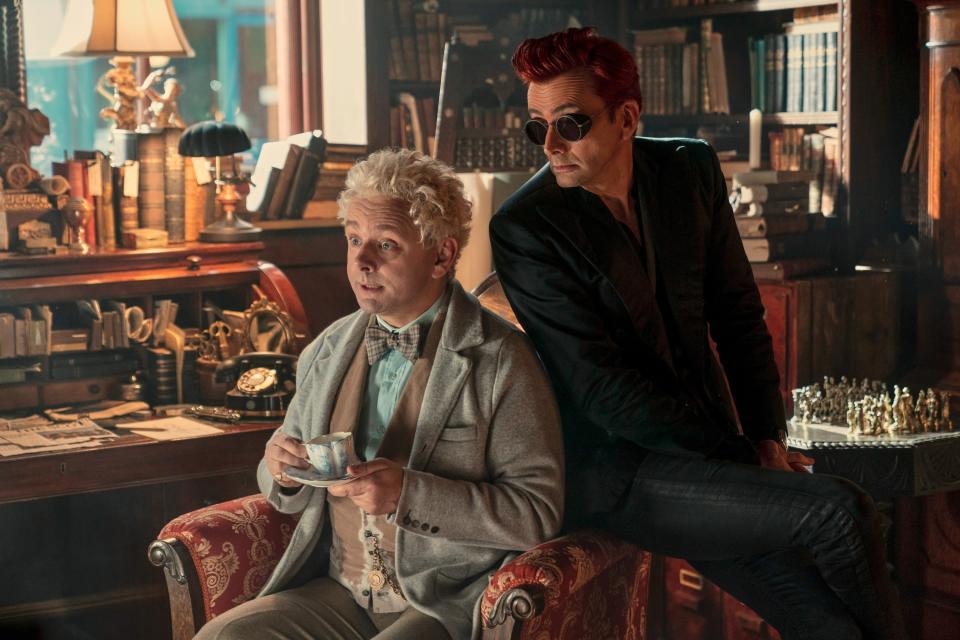 Michael Sheen and David Tennant in 'Good Omens'. Sheen's character sits with a teacup in a classic suit, while Tennant's character, in a dark outfit and sunglasses, leans stylishly