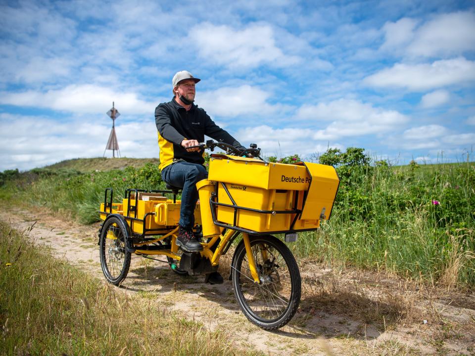 A man in jeans and a black and yellow short ride a tricycle with large cargo baskets on the front and back through a field.